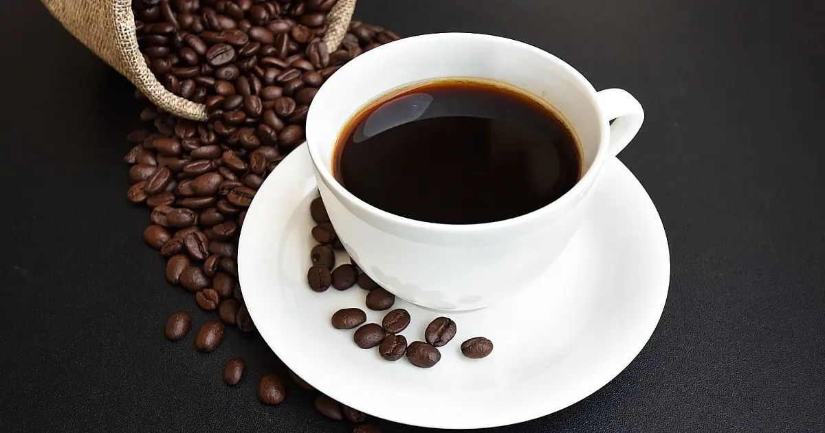 Uma xícara de porcelana preta cheia de café expresso quente, posicionada sobre uma mesa de madeira escura cercada por grãos torrados de café espalhados ao redor