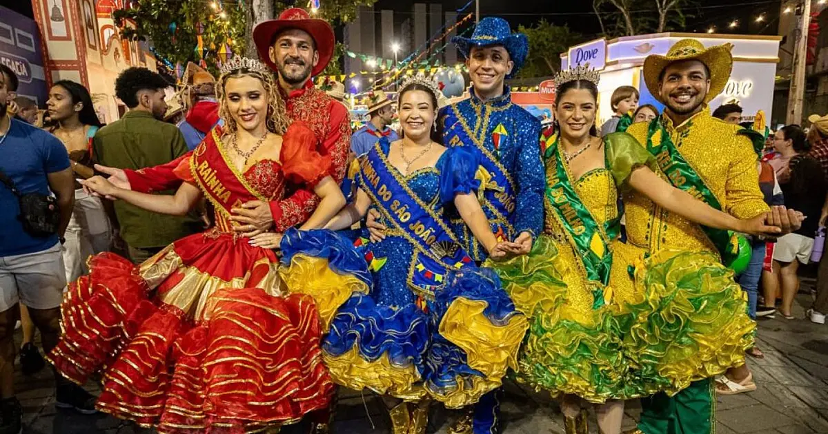 Casais integrantes de uma quadrilha junina tradicional, vestindo roupas matutas coloridas de chita e chapéus de palha, dançando sorridentes em meio ao público na Estação Ferroviária iluminada.