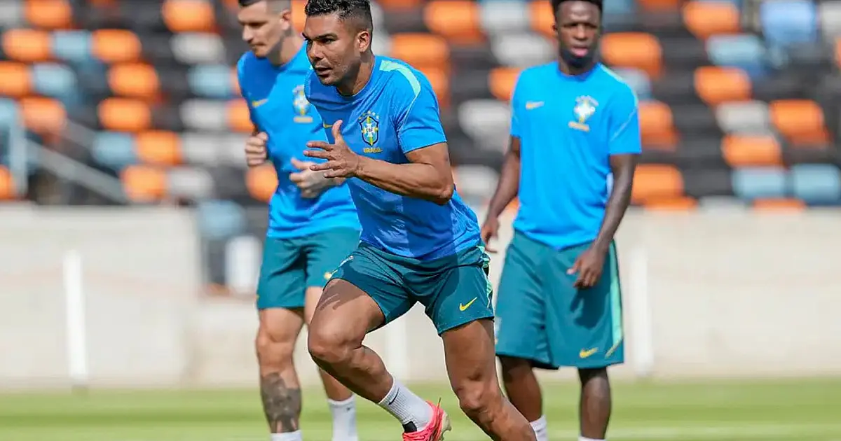 Grupo de atletas com coletes verdes e camisas azuis de treino correndo em fila sobre o gramado verde de um estádio, sob a luz do dia