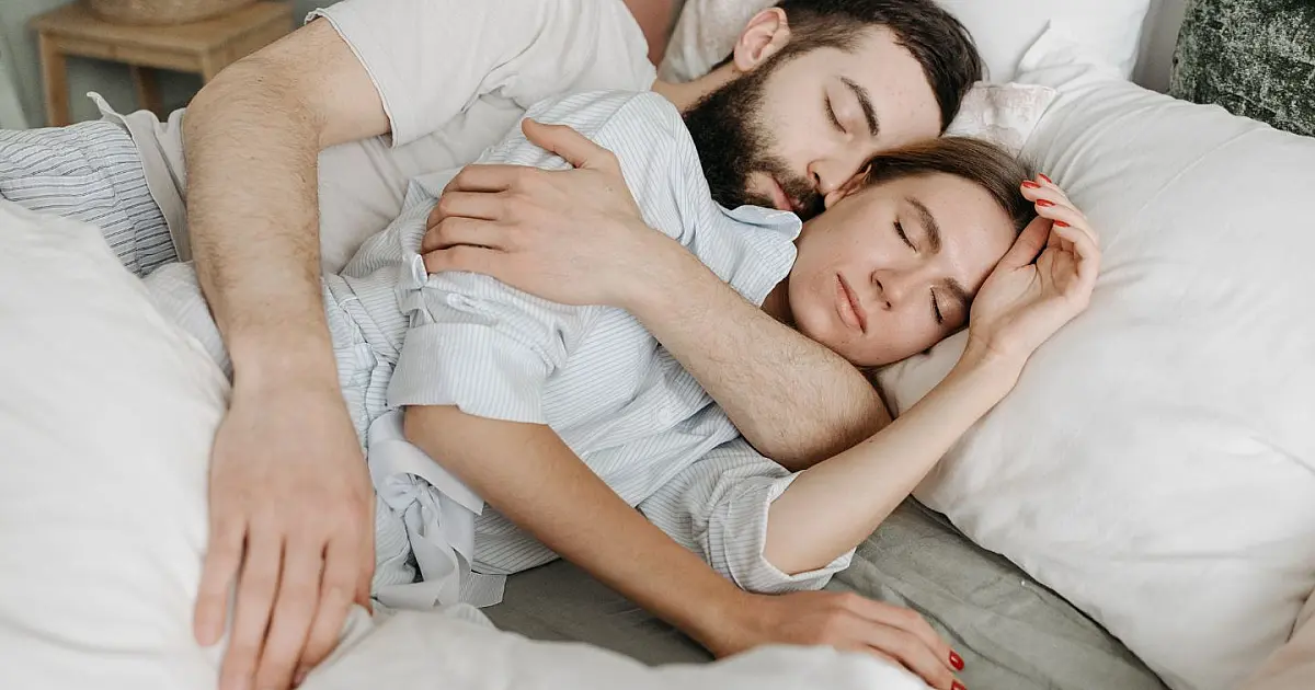Imagem em plano médio de um casal deitado sob lençóis brancos em uma cama confortável, vistos de costas, dormindo abraçados na posição de conchinha