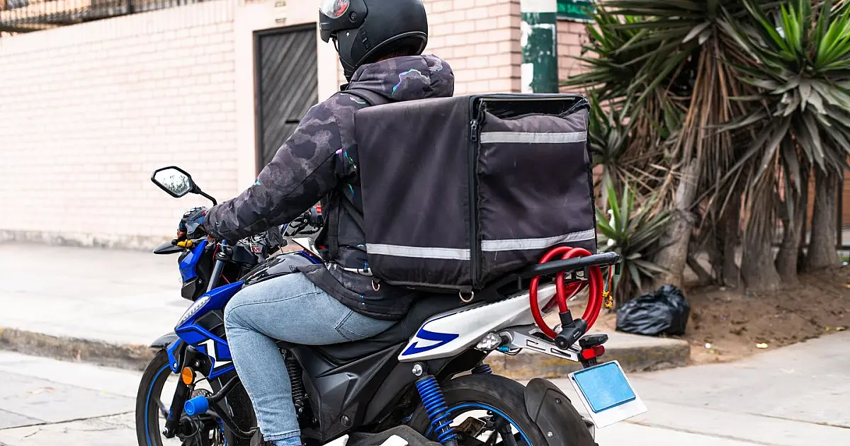 Entregador de aplicativo pilotando uma moto em uma via urbana durante o dia, carregando uma mochila térmica nas costas