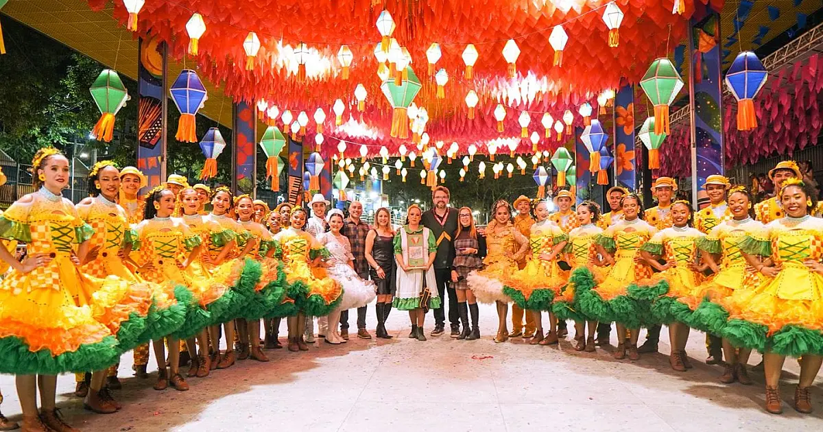 Integrantes de uma quadrilha junina caracterizados com roupas coloridas comemorando e sorrindo após apresentação em um ginásio enfeitado