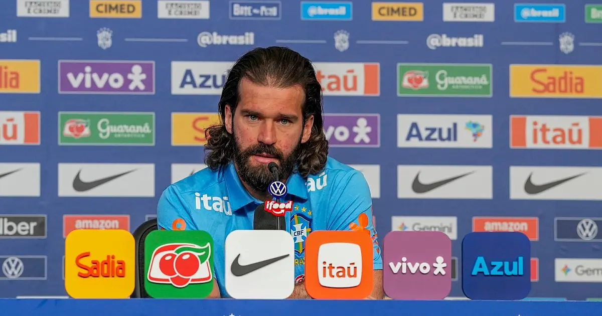 O goleiro Alisson, com uniforme de treino da Seleção Brasileira, falando ao microfone em uma bancada de entrevista coletiva.