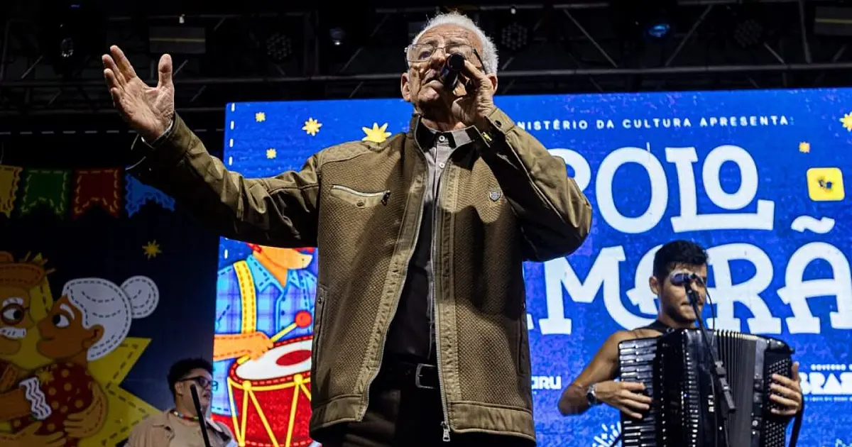 Cantor de forró segurando microfone no palco iluminado da Estação Ferroviária, com público ao fundo vestindo camisas xadrezes