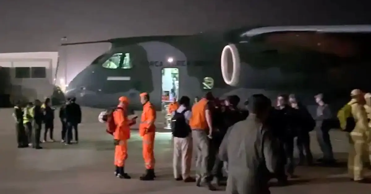 Operários e militares em uma pista de pouso empurrando paletes carregados com caixas identificadas com a bandeira do Brasil em direção à rampa de carga de um avião cinza