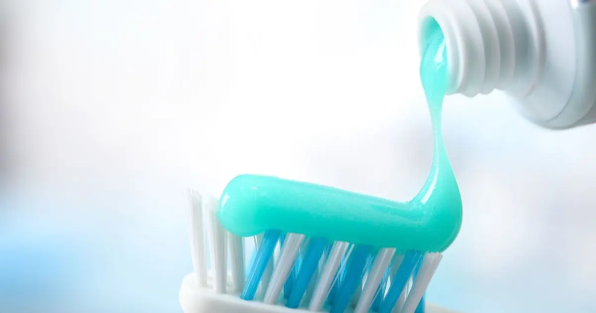 Uma escova de dentes com cerdas macias recebendo uma porção de creme dental branco e consistente, posicionada em cima de uma bancada de banheiro limpa