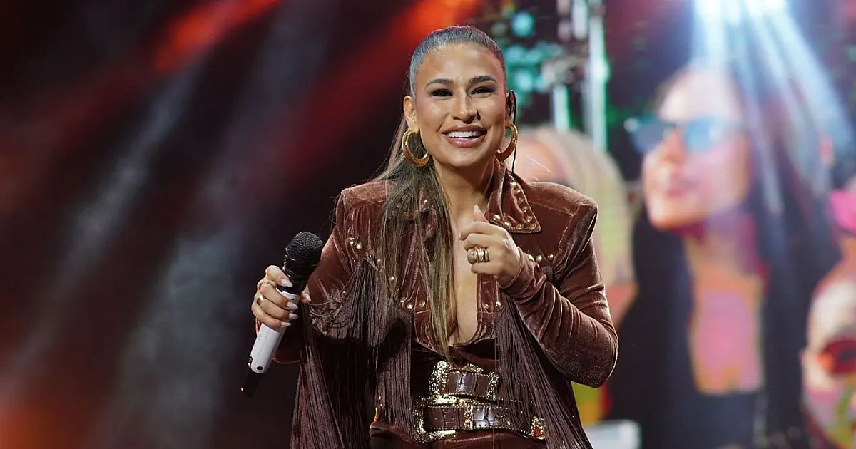 A cantora Simone Mendes sorrindo no palco, vestindo um traje de show brilhante e segurando um microfone sob as luzes coloridas dos refletores
