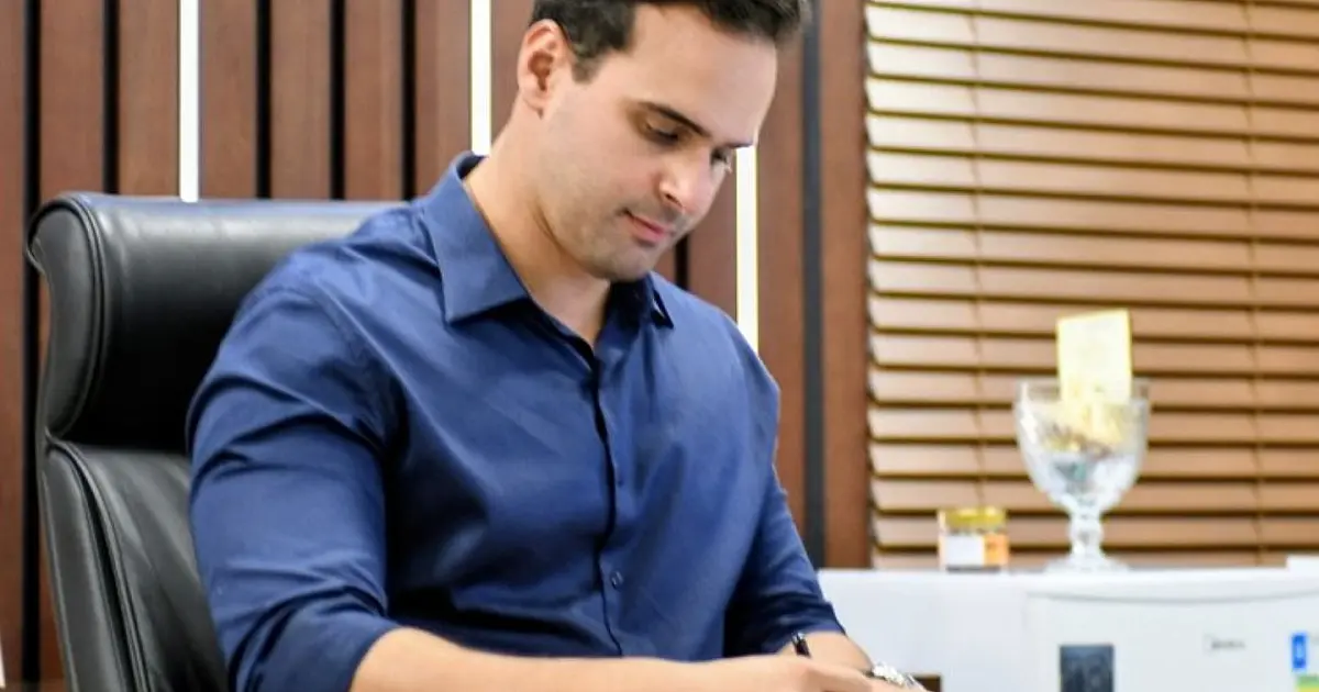 O governador Lucas Ribeiro sentado em sua mesa de gabinete, sorrindo enquanto assina uma série de documentos oficiais com uma caneta preta