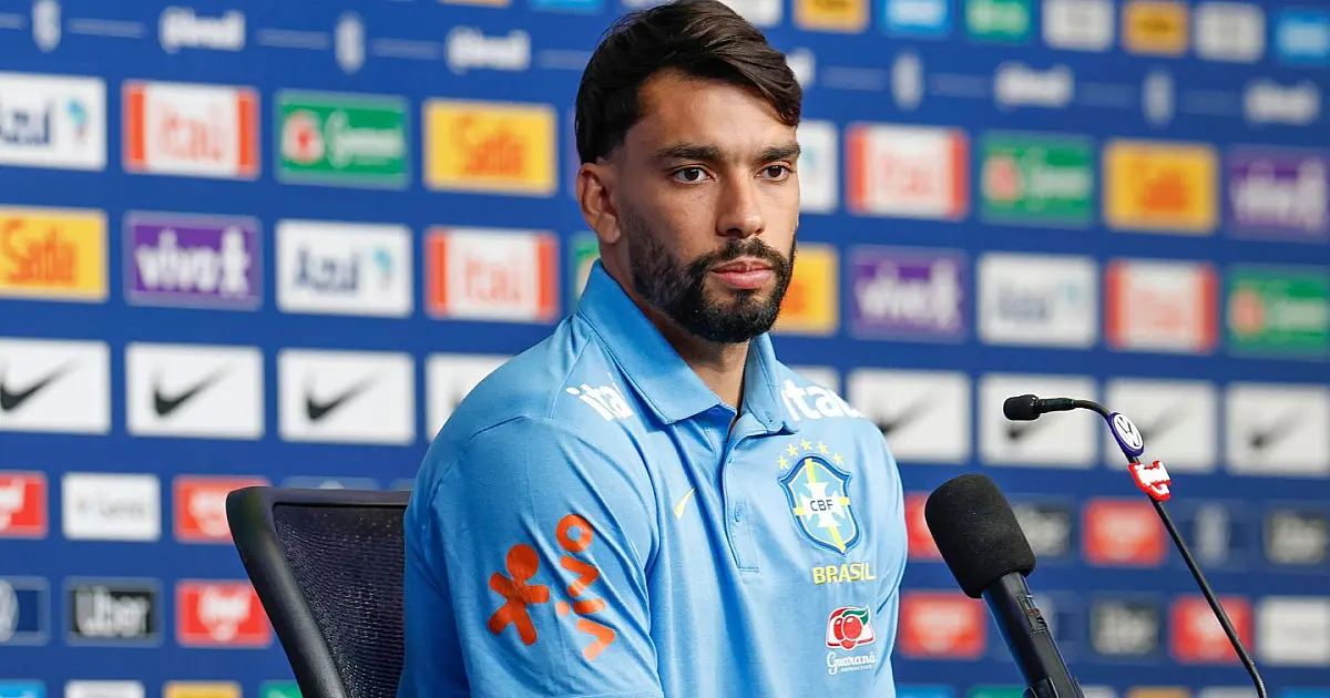 O meio-campista Lucas Paquetá sentado na bancada de entrevistas coletivas, vestindo a camisa de treino azul escura do Brasil, falando ao microfone com banners de patrocinadores ao fundo.