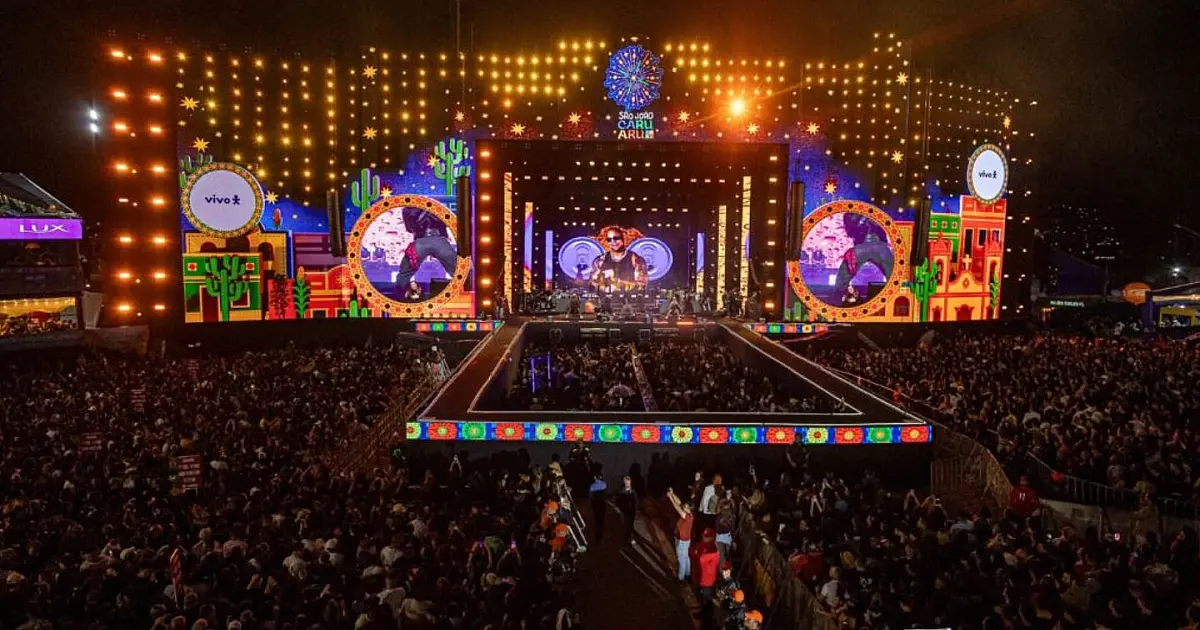 Vista de cima de um palco monumental iluminado por luzes coloridas diante de um mar de pessoas com os braços para cima