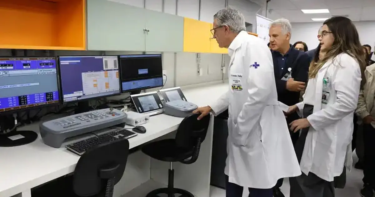 O ministro Alexandre Padilha vestindo terno azul, observando e apontando para uma grande máquina tecnológica de tratamento por radioterapia em uma sala hospitalar moderna