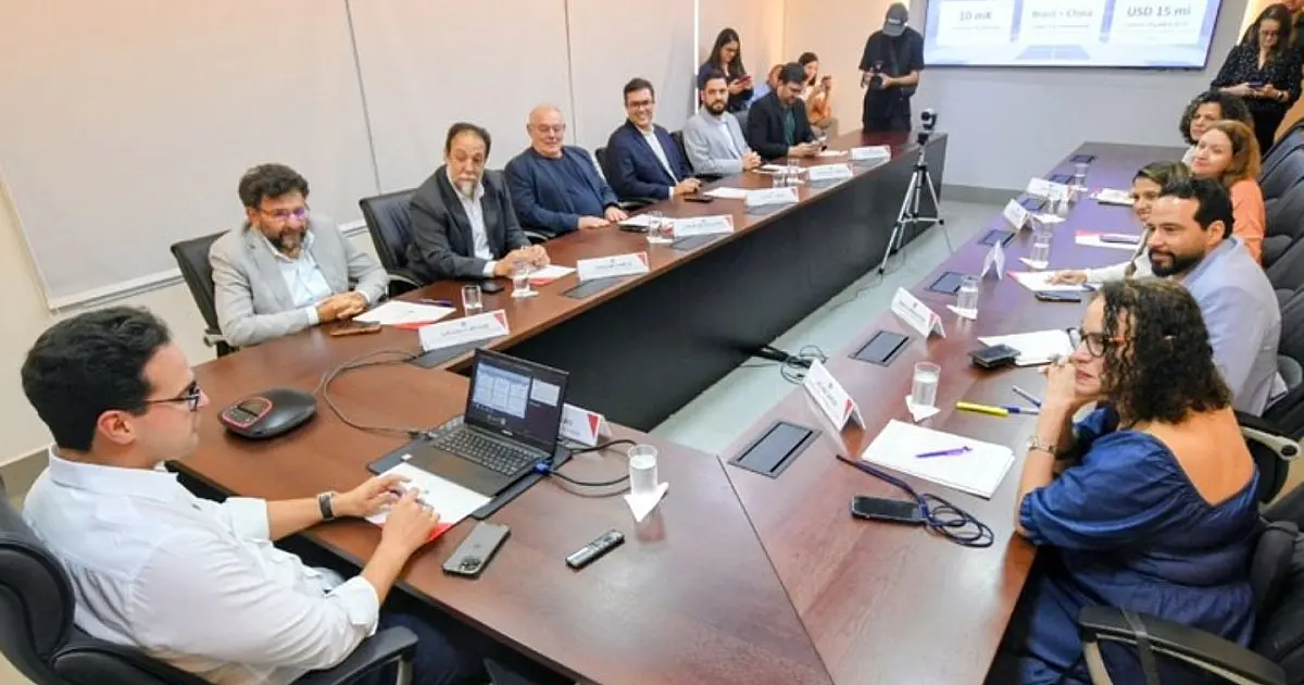 Autoridades e secretários sentados ao redor de uma longa mesa de madeira em uma sala de reuniões formal, ouvindo o governador e a ministra exporem documentos