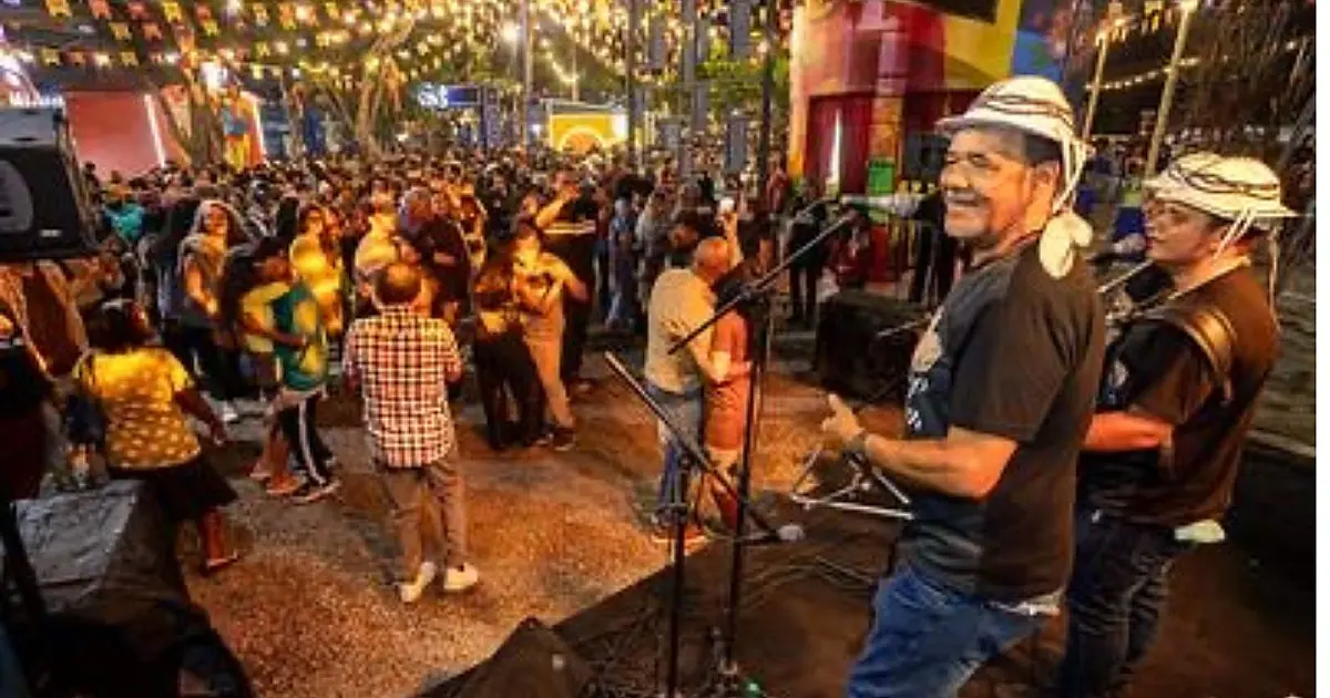 Casal de turistas tirando foto sorridente em frente a uma casinha cenográfica de barro decorada com bandeirinhas juninas