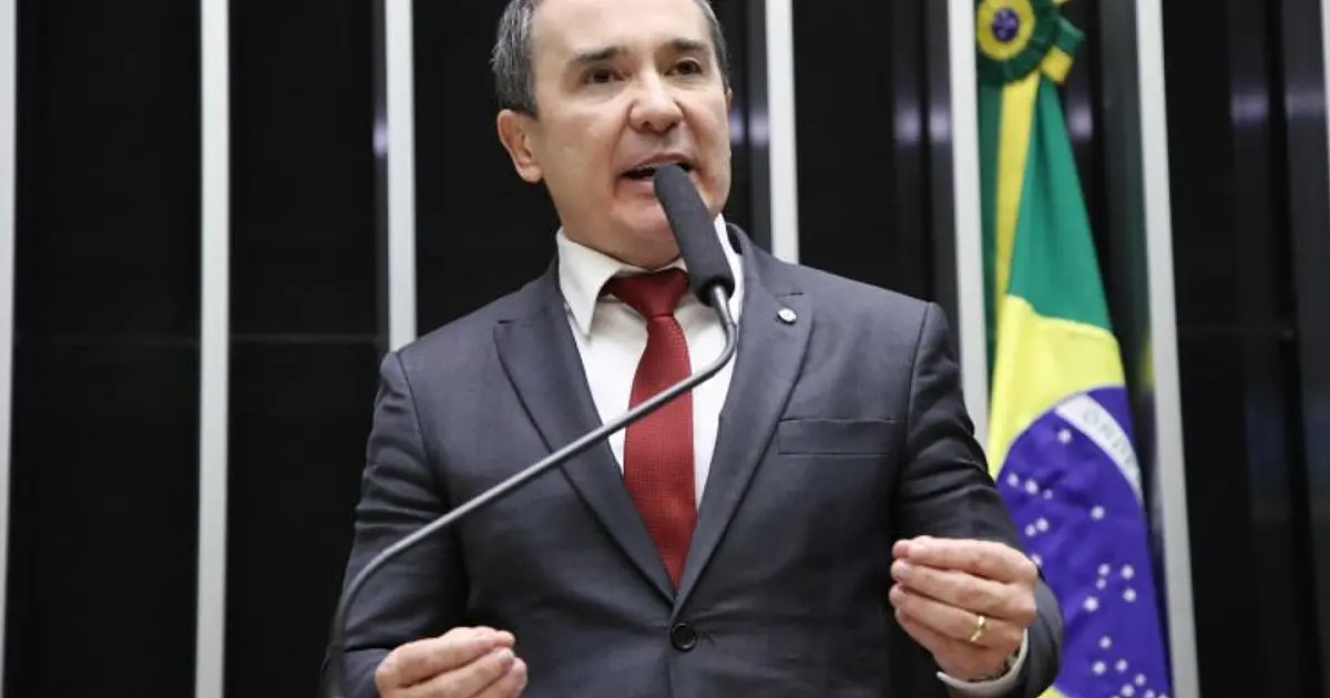 Vista interna de uma sala de comissões na Câmara dos Deputados, com parlamentares sentados ao redor de uma grande mesa de debates de madeira e microfones posicionados.