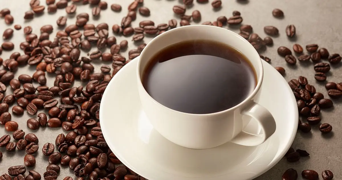 Xícara de porcelana branca cheia de café preto fumegante apoiada em uma mesa de madeira escura e cercada por grãos torrados