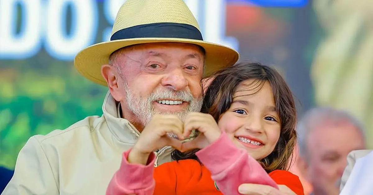 O presidente Luiz Inácio Lula da Silva sorrindo ao lado de uma menina em um evento público oficial, cercado por faixas e bandeiras do programa habitacional e de reforma agrária