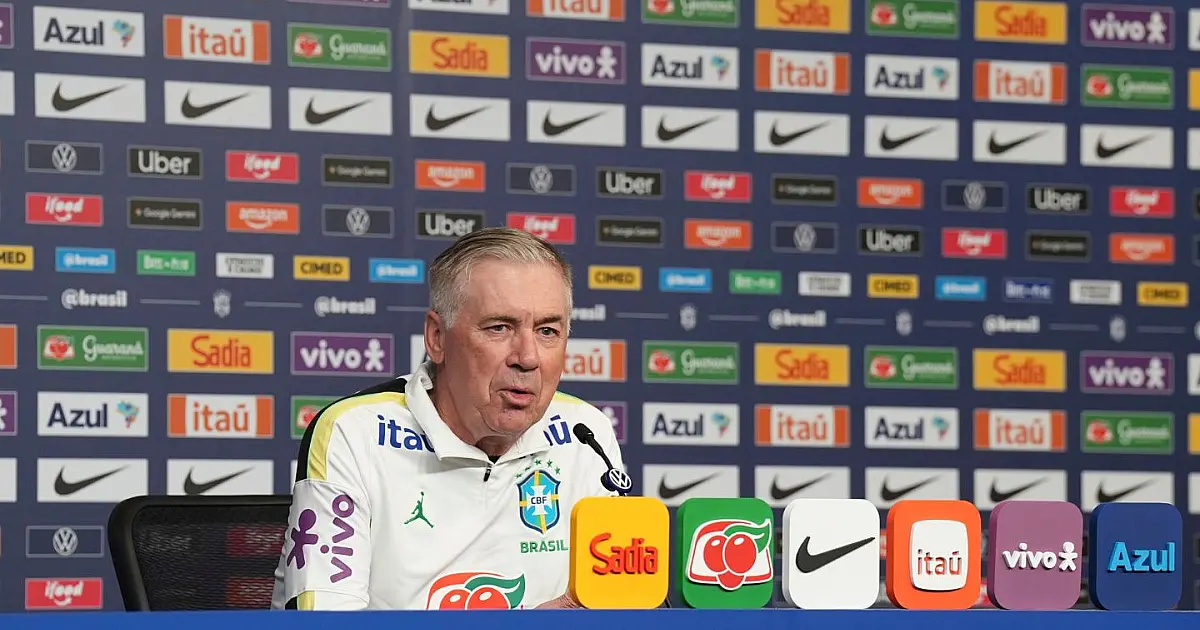 Técnico Carlo Ancelotti sentado em uma bancada de coletiva de imprensa falando ao microfone com o painel de patrocinadores ao fundo