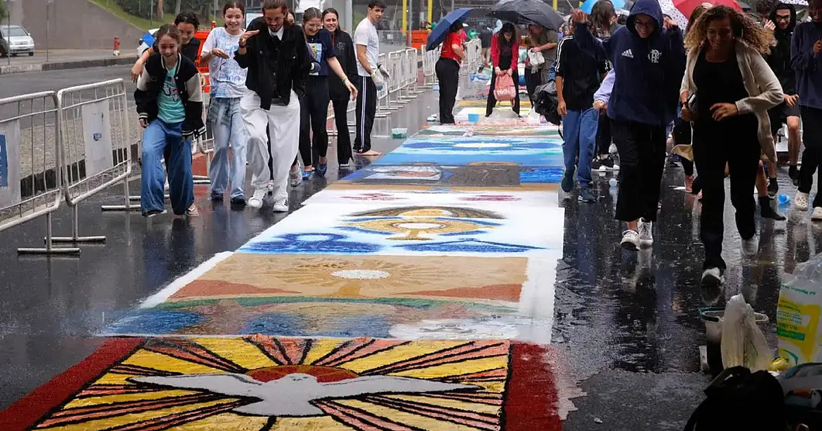 Pessoas de joelhos sobre o asfalto espalhando sal colorido para moldar um grande tapete decorativo em frente a uma igreja