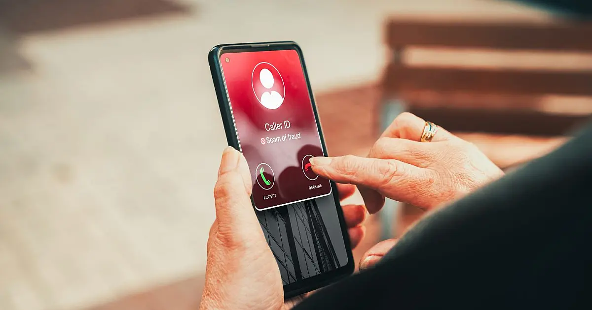 Mão de uma pessoa idosa segurando um telefone celular que exibe um alerta de chamada suspeita na tela de toque, com o dedo indicador prestes a recusar a ligação