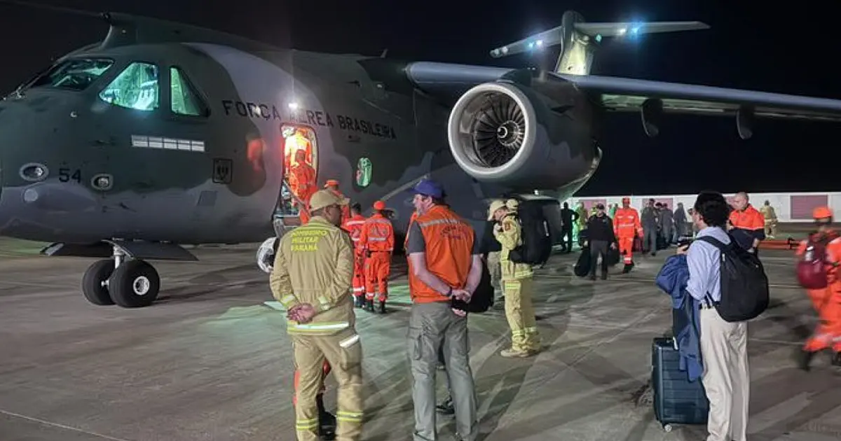 Bombeiros militares brasileiros vestindo fardamento laranja e capacete tático, acompanhados de um cão farejador, inspecionando uma pilha de blocos de concreto e destroços de um prédio desabado