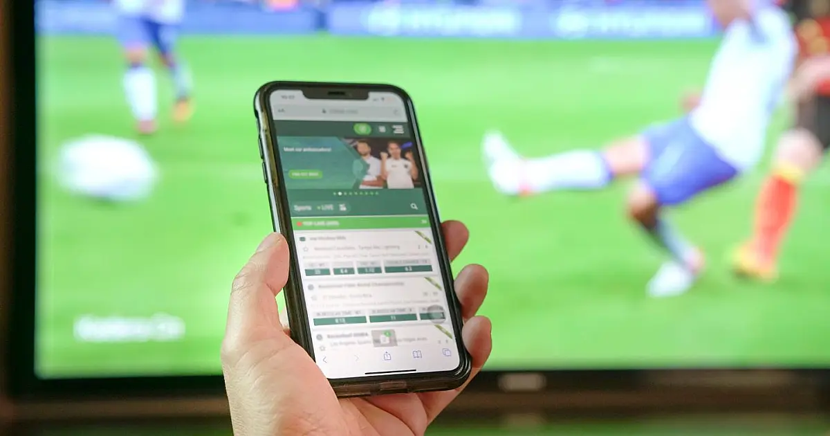 close-up de uma pessoa segurando um smartphone com as duas mãos, mostrando um aplicativo com gráficos de apostas esportivas na tela, contra um fundo escuro desfocado