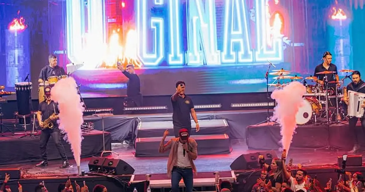O cantor Zé Vaqueiro sorrindo e cantando no microfone durante um show noturno com efeitos visuais ao fundo