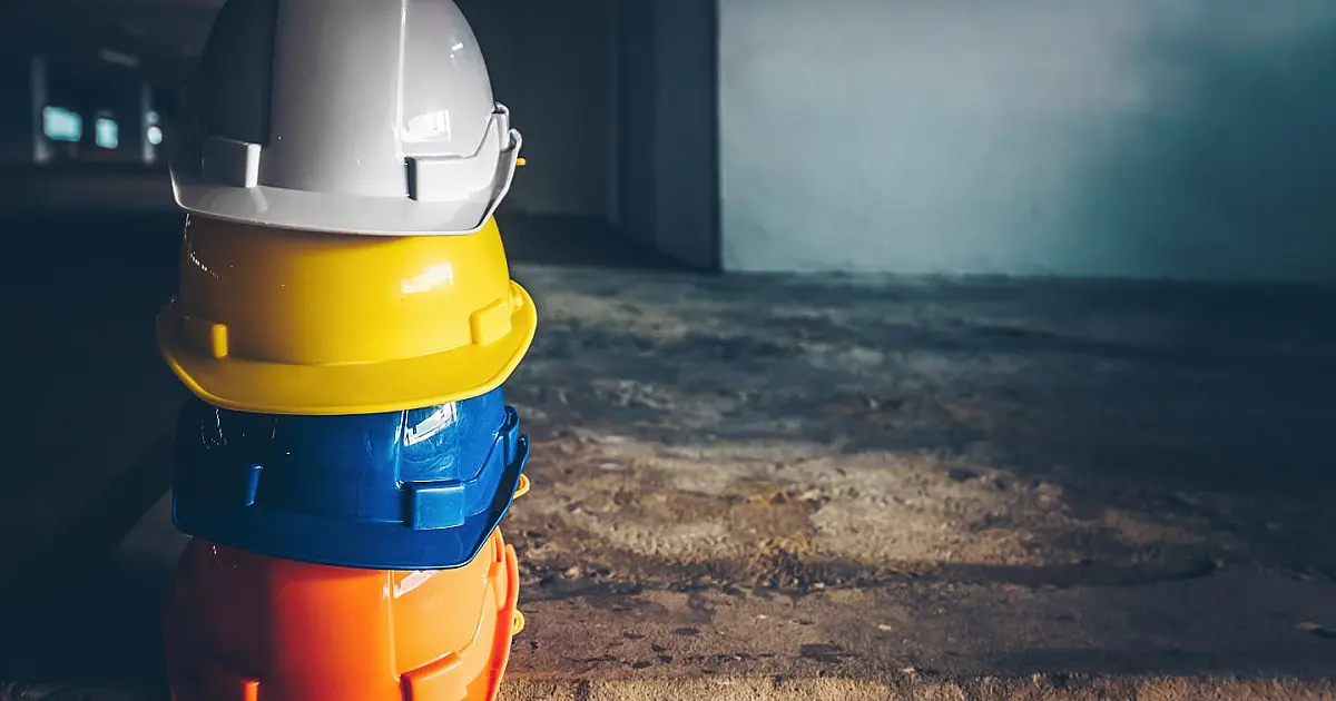 Linha horizontal exibindo capacetes de proteção novos nas cores branca, amarela, azul, verde e vermelha alinhados sobre uma viga de concreto