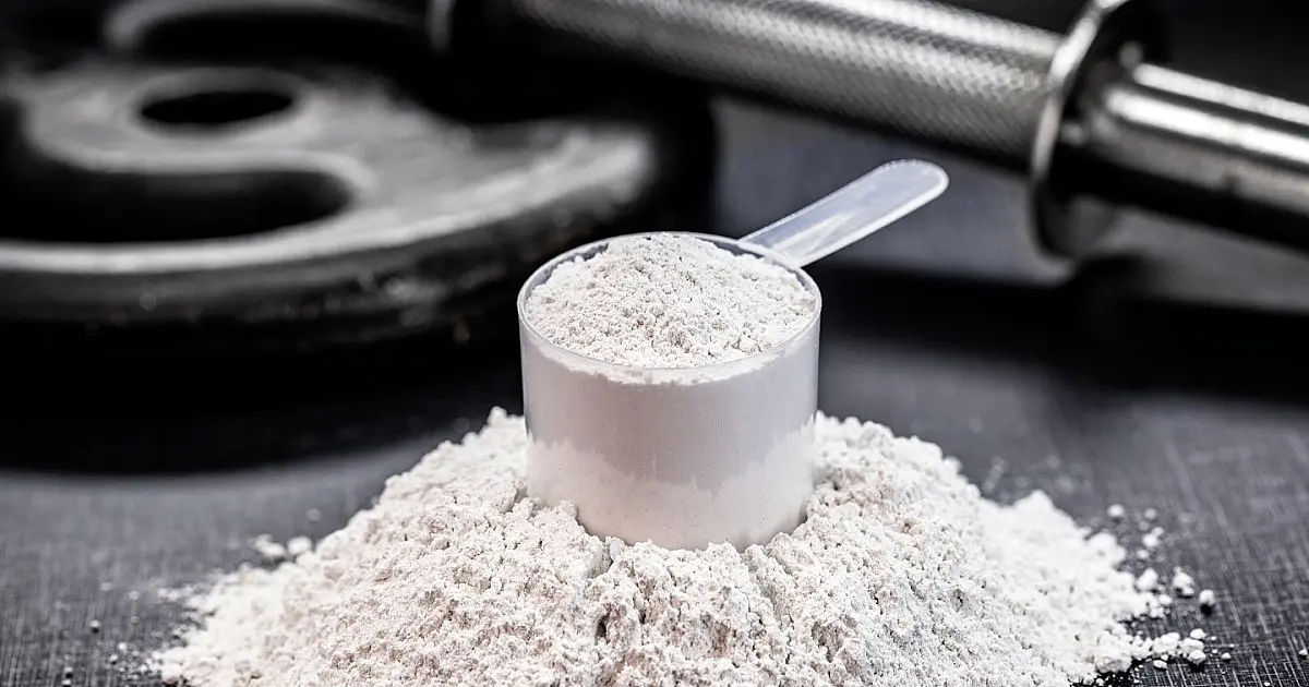 Colher de medida contendo suplemento em pó branco posicionado ao lado de potes de nutrição esportiva